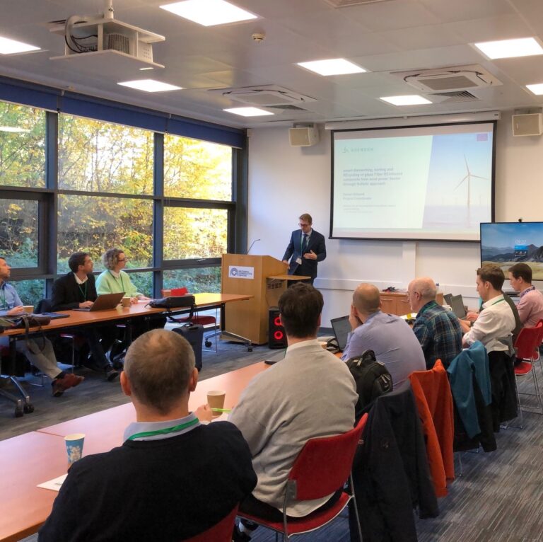 A man stands at the front of a room giving a presentation to a group of people seated at tables.