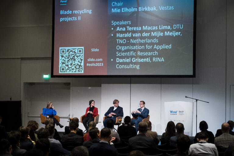 4 people seated in front of an audience. There is a large screen on the wall behind them.