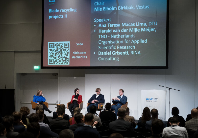 4 people seated in front of an audience. There is a large screen on the wall behind them.