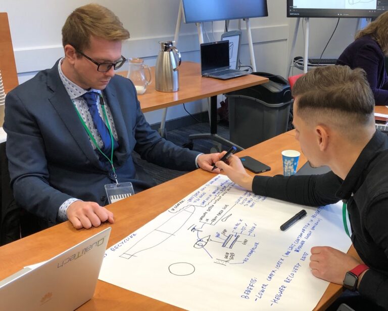 Two people seated at a table drawing a diagram on a large sheet of paper.