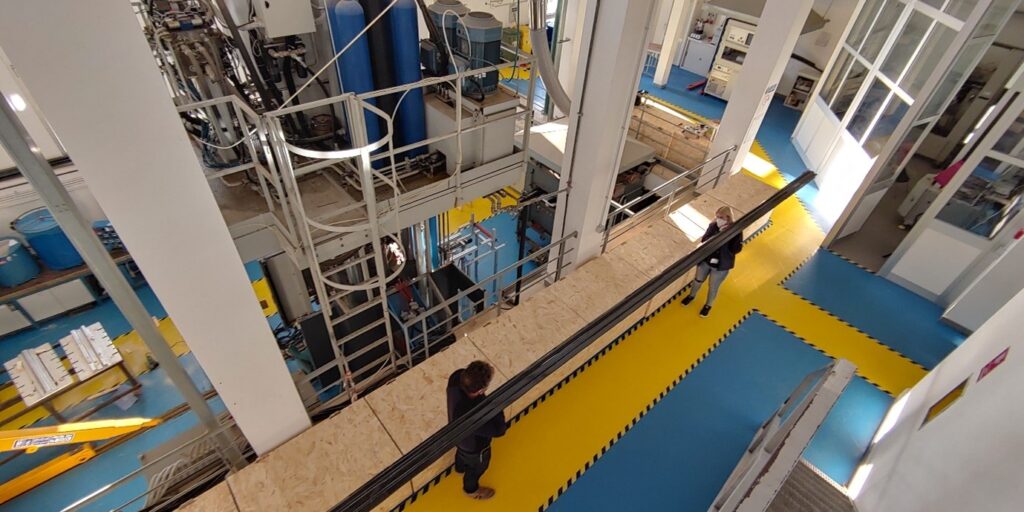 Two people holdng a long pole, standing in a hall full of industial equipment.