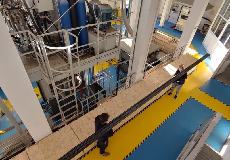 Two people holdng a long pole, standing in a hall full of industial equipment.