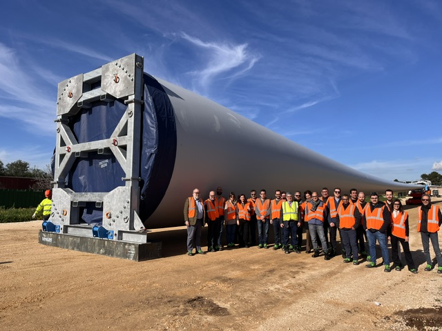 Group of people in high-vis jackets stand at side of the root of a wind turbine blade.