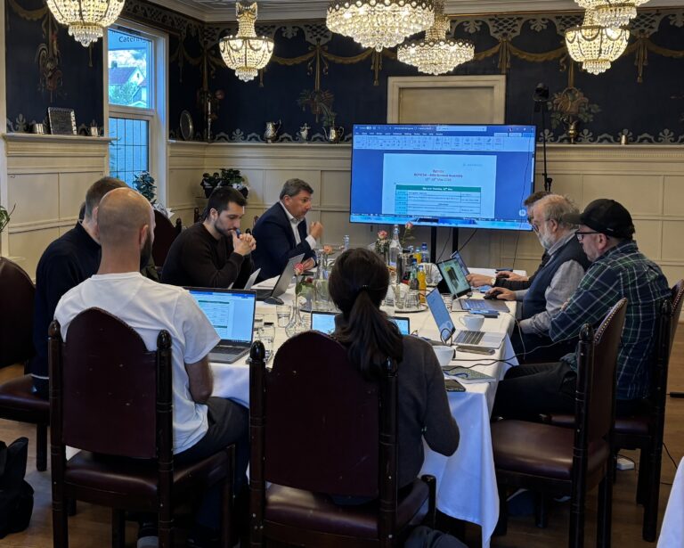 Meeting A group of people seated at a table in front of a large screen.